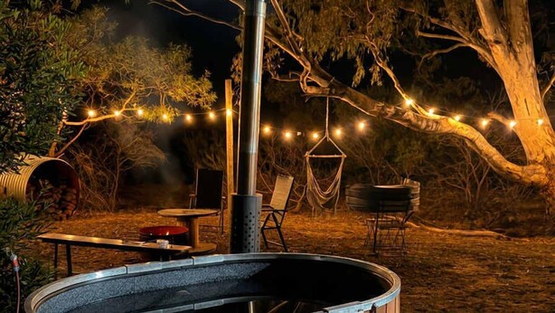 A hot tub with a wooden exterior, surrounded by outdoor furniture and string lights.