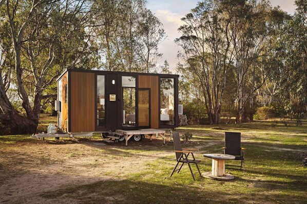 Una cabaña moderna y compacta con fachada de madera y ventanas amplias, ubicada en zona boscosa con una mesa y sillas de picnic cerca.