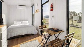 A bedroom with a bed, a table, and chairs, a window with a view of the outdoors, and a fire extinguisher mounted on the wall. - Rosebrook Riverviews by Tiny Away (Rosebrook)
