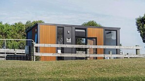 A modern, compact building with a wooden exterior and large windows, surrounded by a grassy area and a wooden fence. - Rosebrook Riverviews by Tiny Away (Rosebrook)