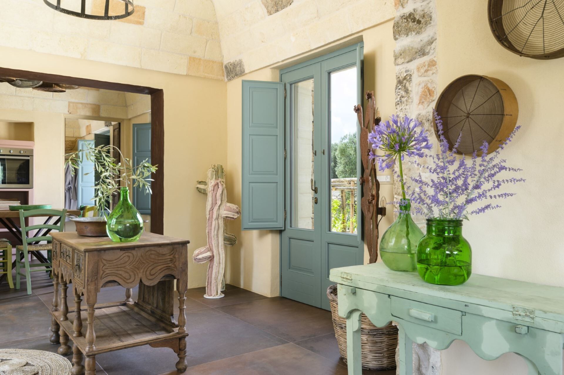 Un interior rústico con una mesa de madera, un jarrón verde con flores, una puerta azul y una pared de piedra.