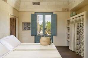 A bedroom with a bed, a window with open shutters, a woven basket, and a shelf with decorative items.