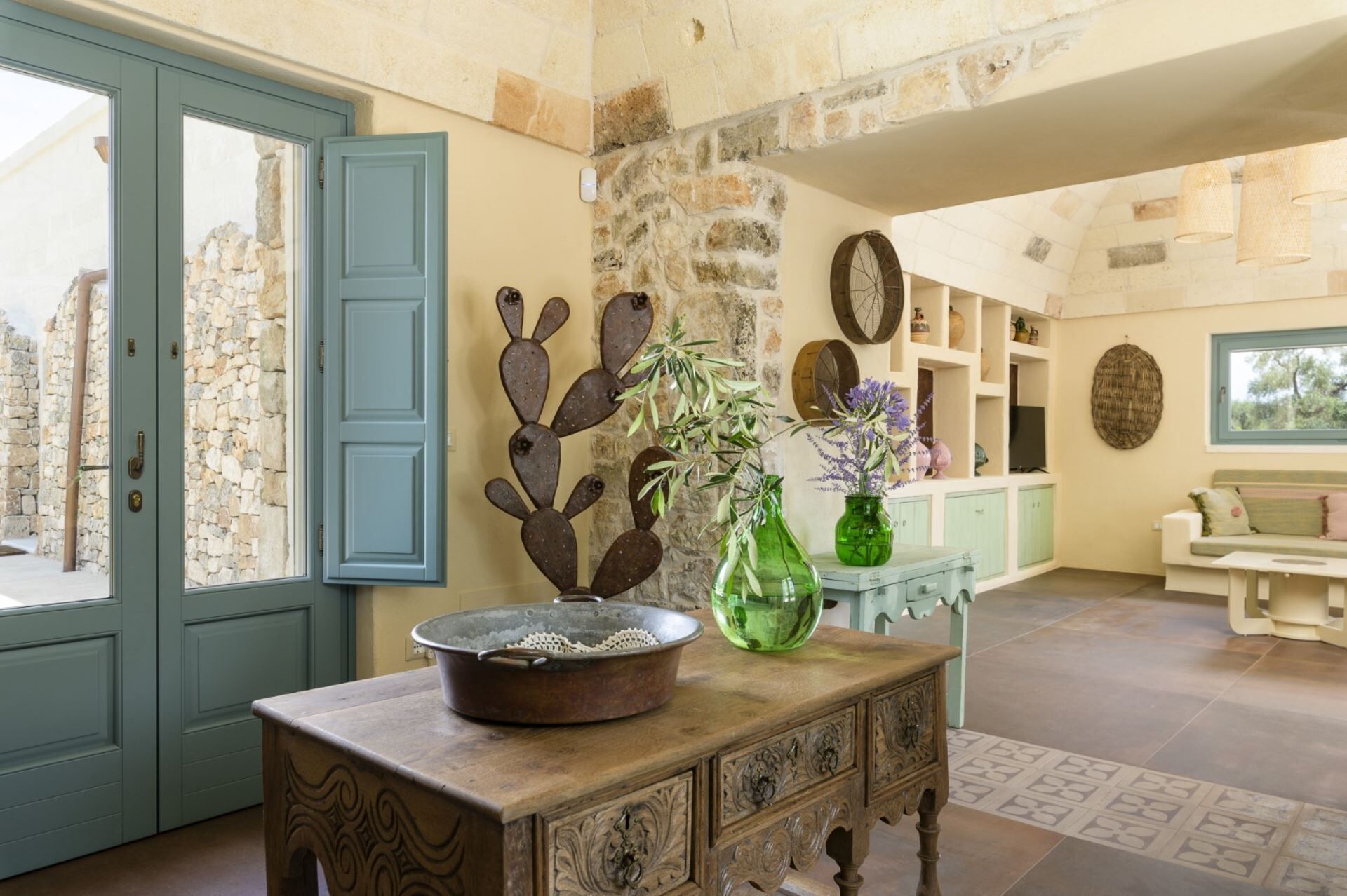 Una habitación con pared de piedra, una puerta azul, una mesa de madera rústica y un jarrón verde con flores.