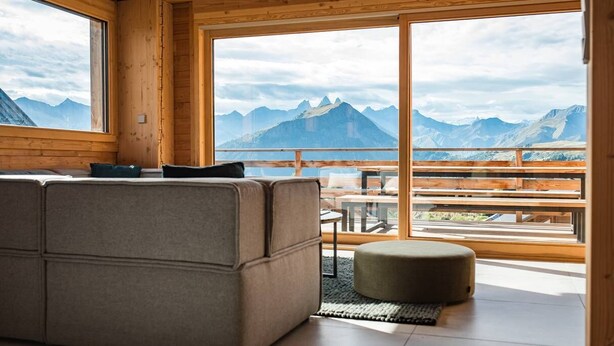 A modern log cabin interior with a sofa, ottoman, and large windows offering mountain views. - Semi-detached chalet near the slopes with sauna (LA TOUSSUIRE)