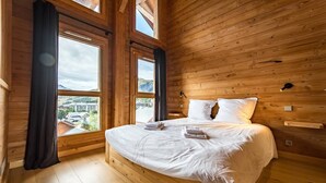 A wooden cabin bedroom with a large bed, two windows, and a view of a town. - Semi-detached chalet near the slopes with sauna (LA TOUSSUIRE)