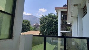 A view from a window with a balcony, overlooking a building with a green roof, trees, and a mountain in the background. - HIGHLAND BREEZE AVACADO HOTEL (Mbeya)