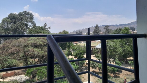 A balcony with a metal railing overlooking a landscape with trees and a hill in the distance. - HIGHLAND BREEZE AVACADO HOTEL (Mbeya)