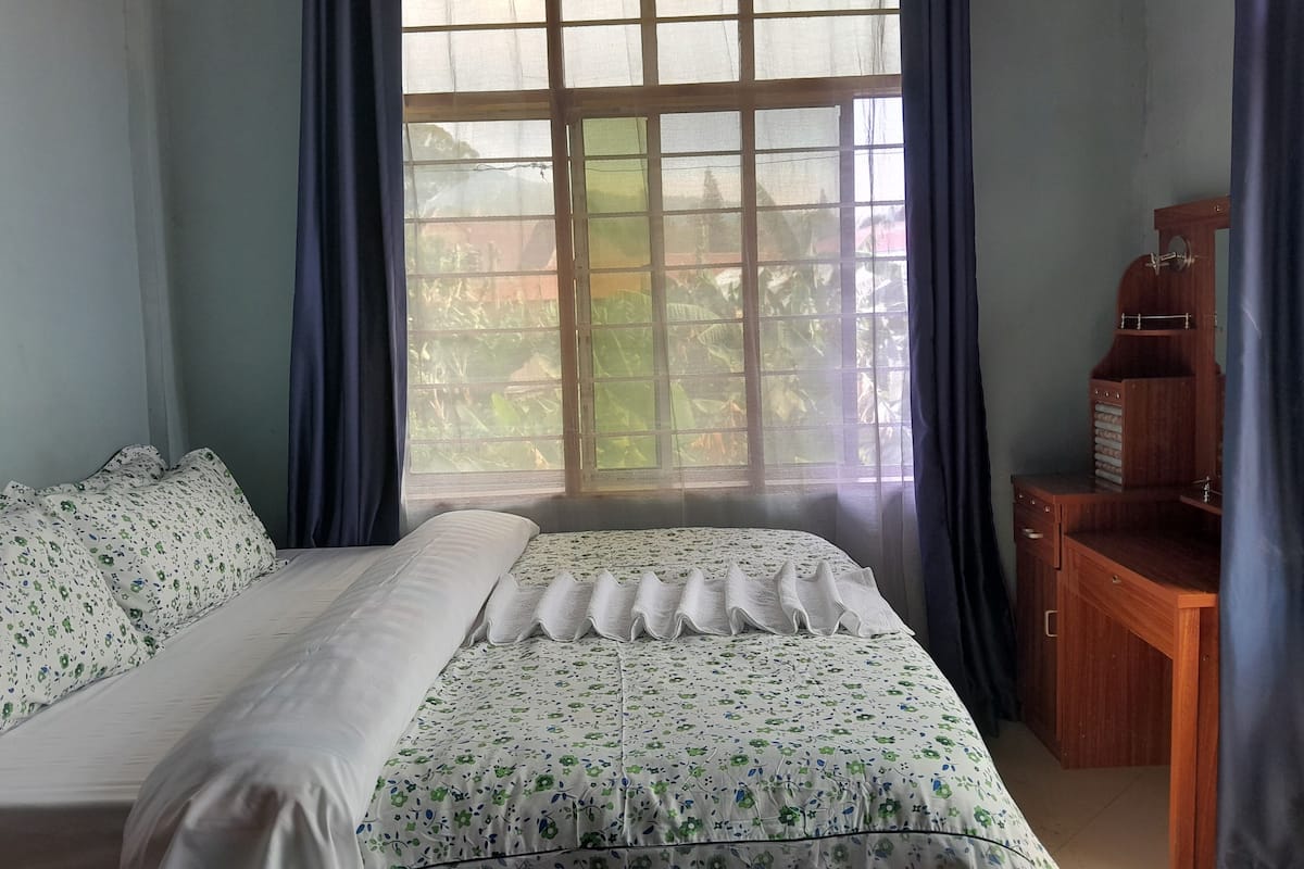 Quarto com cama, escrivaninha de madeira e janela com cortinas.