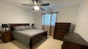 Un dormitorio con cama, mesitas de noche, cómoda, ventilador de techo y ventana con cortinas.