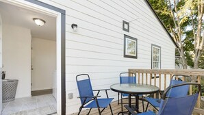 An outdoor patio with a round table and blue chairs, a wooden deck, and a white exterior wall with a door leading inside.