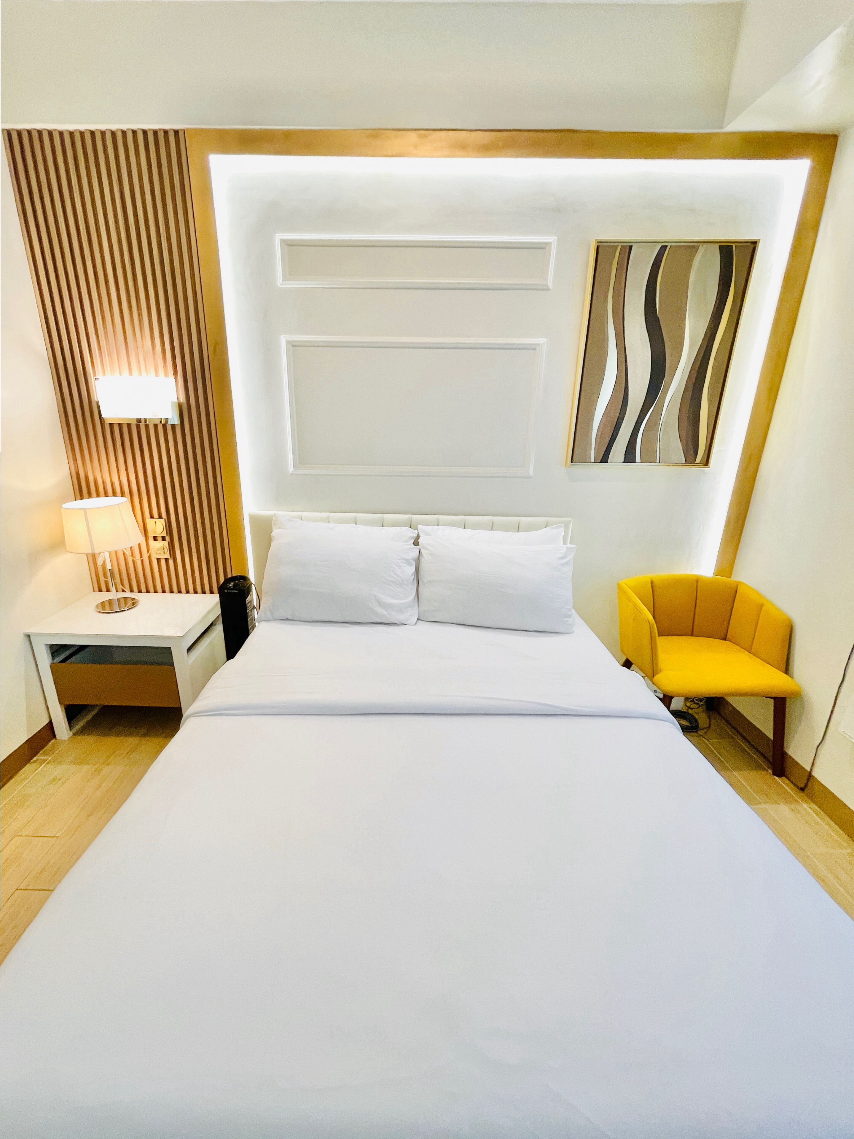 Quarto de hotel com uma cama grande, uma mesinha de cabeceira com luminária, uma cadeira e um ponto de luz fixado na parede.