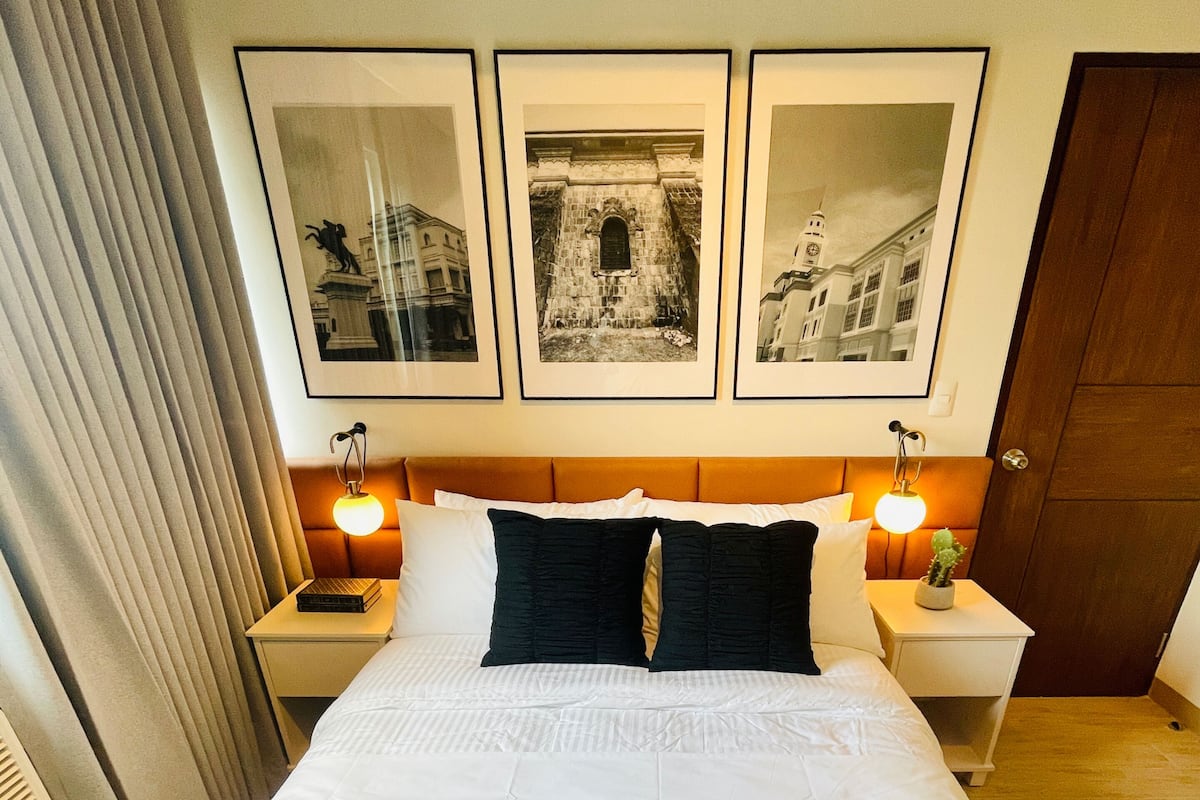 Un lit bien fait, avec du linge de lit blanc, flanqué de deux tables de chevet sur lesquelles reposent des lampes, et trois tableaux encadrés accrochés au mur au-dessus.