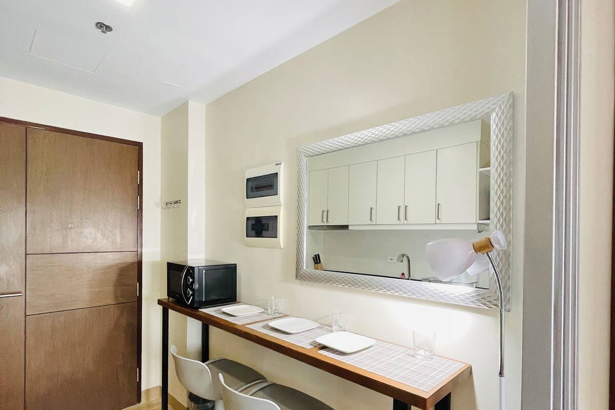 Une chambre d’hôtel compacte dotée d’une kitchenette, d’un coin repas avec deux chaises, d’un grand miroir et d’une armoire en bois.