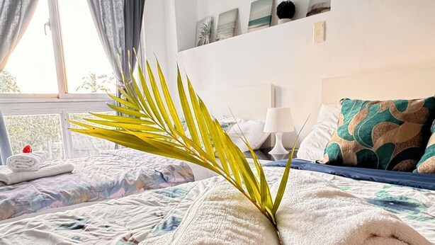 Ein ordentlich bezogenes Bett mit aufgerollten Handtüchern und einem Palmblatt, ein Fenster mit Vorhängen und eine Wand mit gerahmten Kunstwerken.