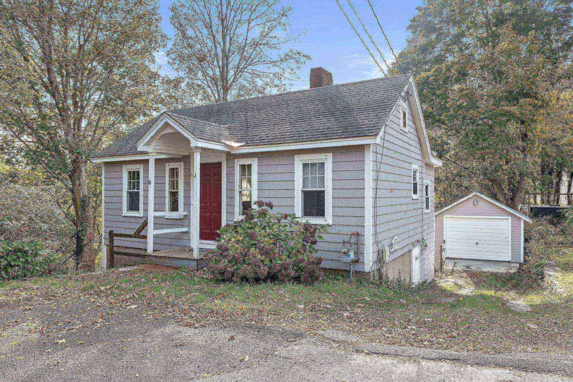 Une petite maison à un étage, avec une porte rouge et des boiseries blanches, entourée d’arbres et adossée à un garage.
