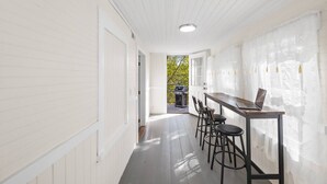 Un couloir étroit aux murs blancs et au plancher en bois, qui mène à un patio équipé d’un barbecue et de mobilier d’extérieur.