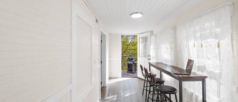 A narrow hallway with white walls and a wooden floor, leading to a patio area with a grill and outdoor seating.