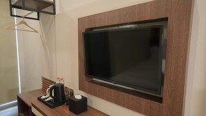 A modern hotel room with a large flat-screen TV mounted on a wooden wall, a wooden bench with a tray table, a black coffee maker, and two bottles of water. - Parkside's Hotel Palembang (Palembang)