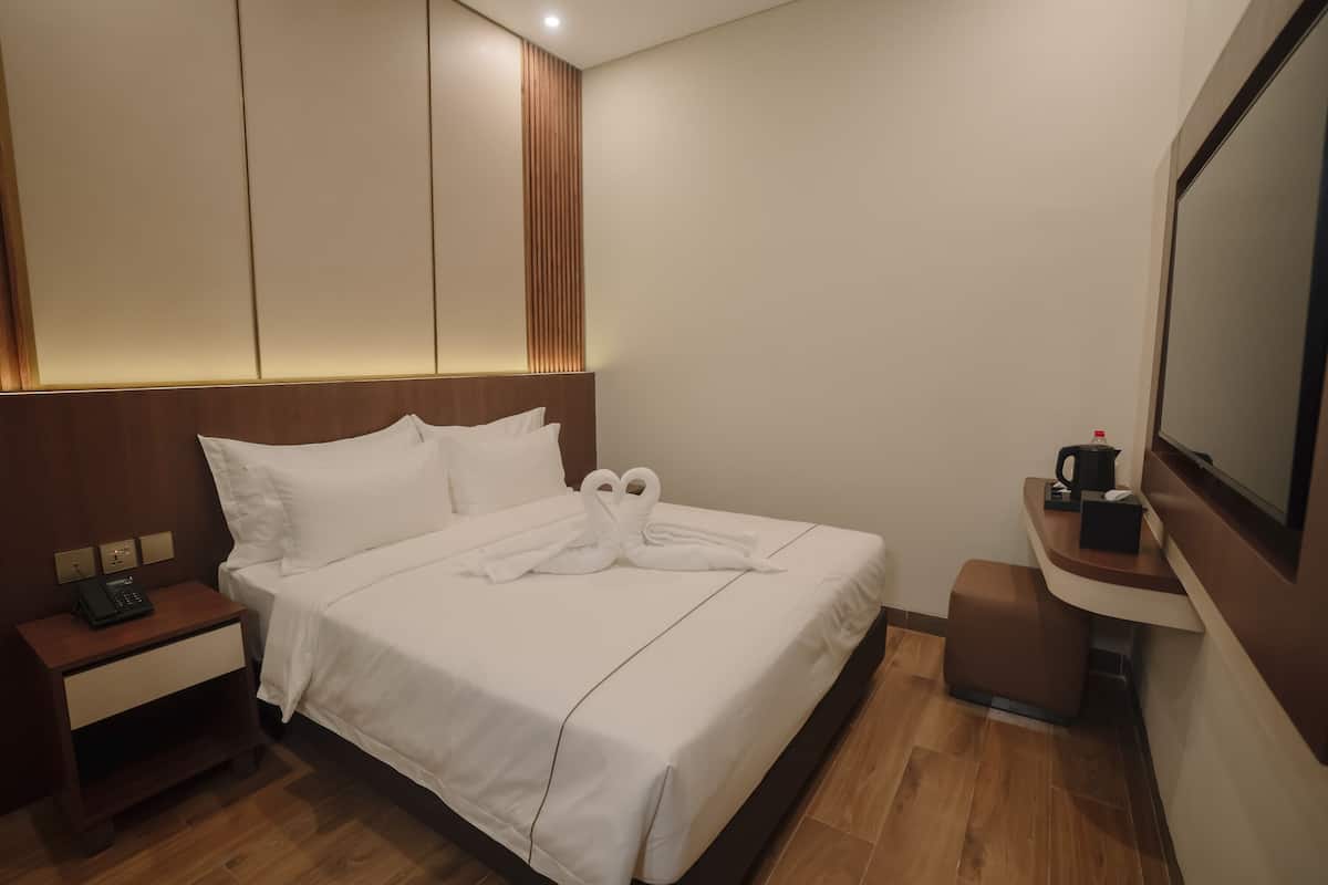 A neatly made bed with white linens and two towels arranged in a heart shape. There is a wooden nightstand with a phone and a small drawer. A coffee maker is on a counter next to a television.
