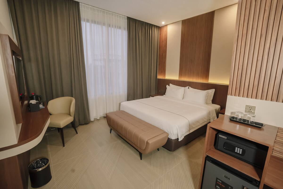 A hotel room with a bed, a desk, a chair, a bench, a trash bin, and a minibar.