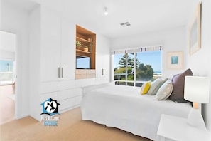 A modern bedroom with a large bed, a built-in TV shelf, and a view of the outdoors.