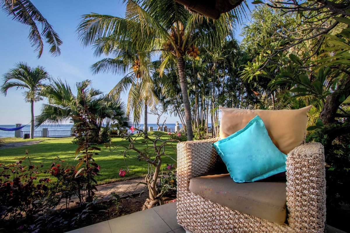A wicker lounge chair with a cushion, positioned in a garden with palm trees and a view of the sea.