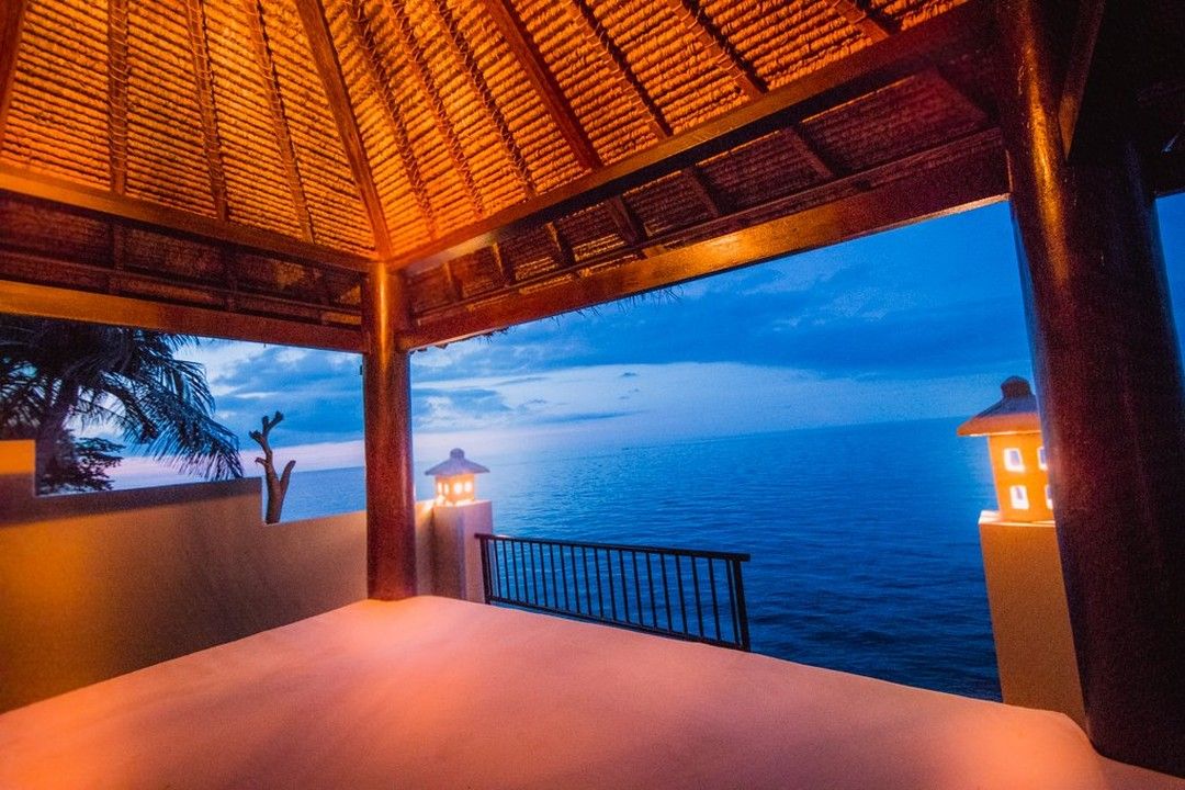 A balcony with a view of the sea at dusk, featuring a thatched roof and two lit lanterns.