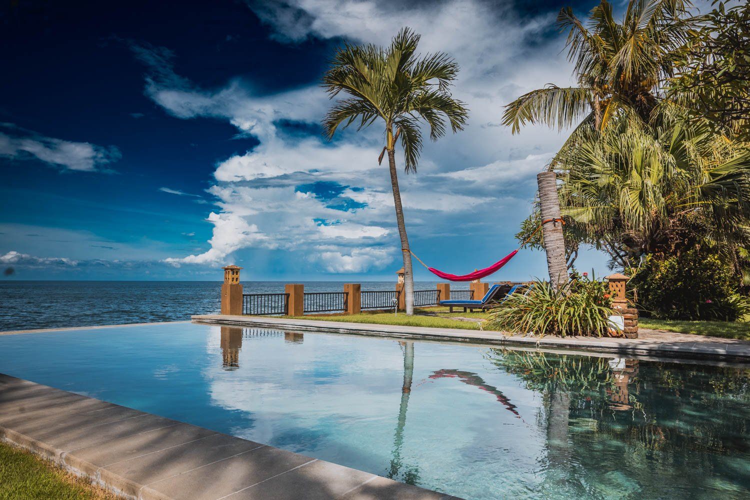A pool with a view of the ocean, palm trees, and a hammock.