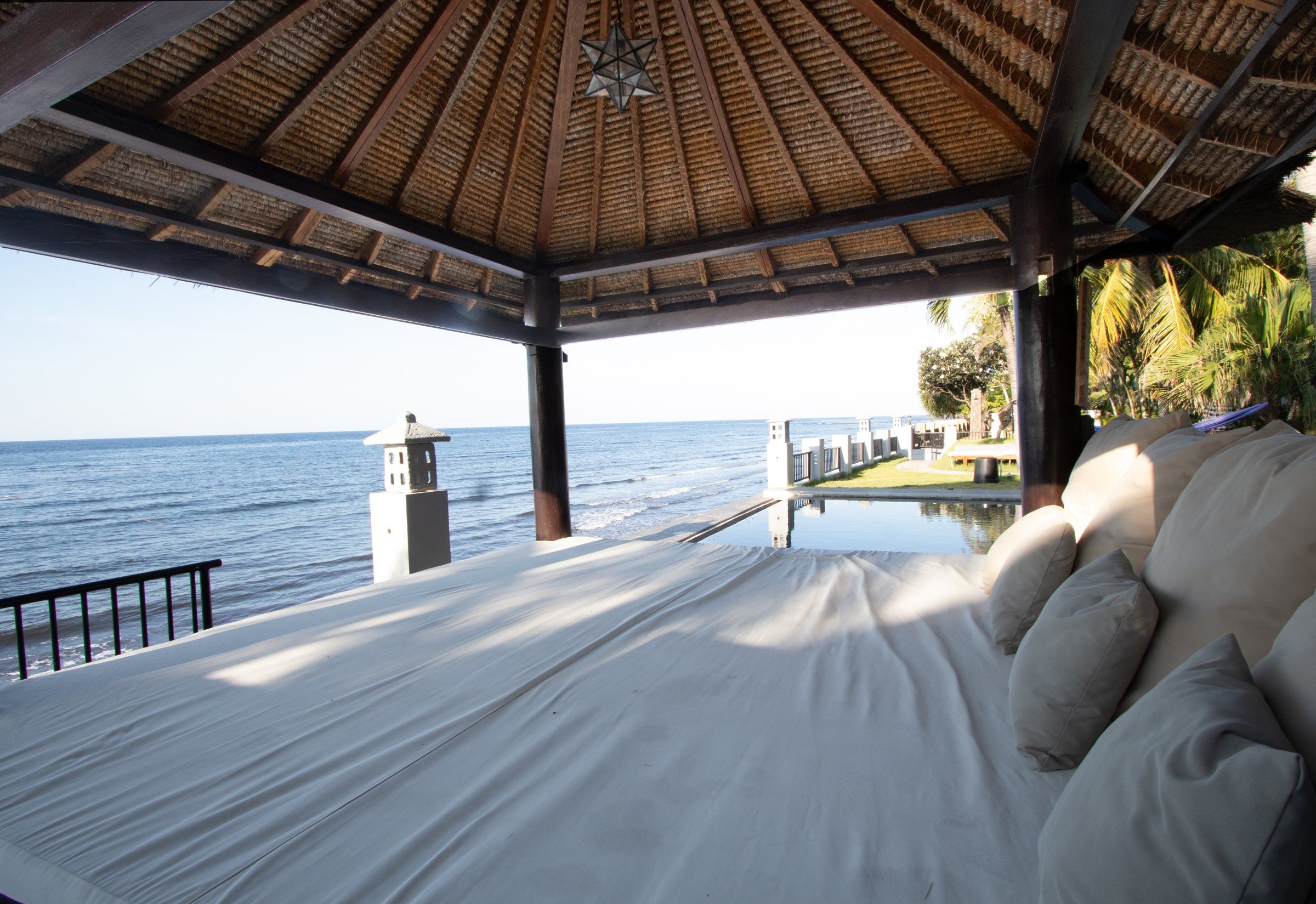 A beachside room with a bed, a view of the sea, a pool, and a thatched roof.