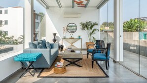 Ein modernes Wohnzimmer mit Sofa, Sessel und Couchtisch, mit einem großen Fenster und Blick ins Freie.