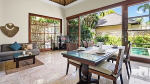 Amplia sala de estar con una mesa de centro de vidrio, un sofá y una mesa de comedor con sillas. Una ventana grande ofrece vistas a una piscina y follaje.