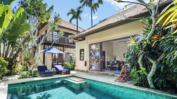 Un área de piscina con sillones de descanso, un espacio de estar cubierto y plantas tropicales.