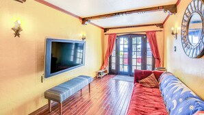 Um quarto aconchegante com piso de madeira, um banco, um sofá com capa estampada, um espelho grande e uma TV fixada na parede.