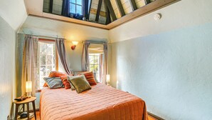 Quarto com cama grande, duas janelas com cortinas, um claraboia e uma pequena mesa com luminária.