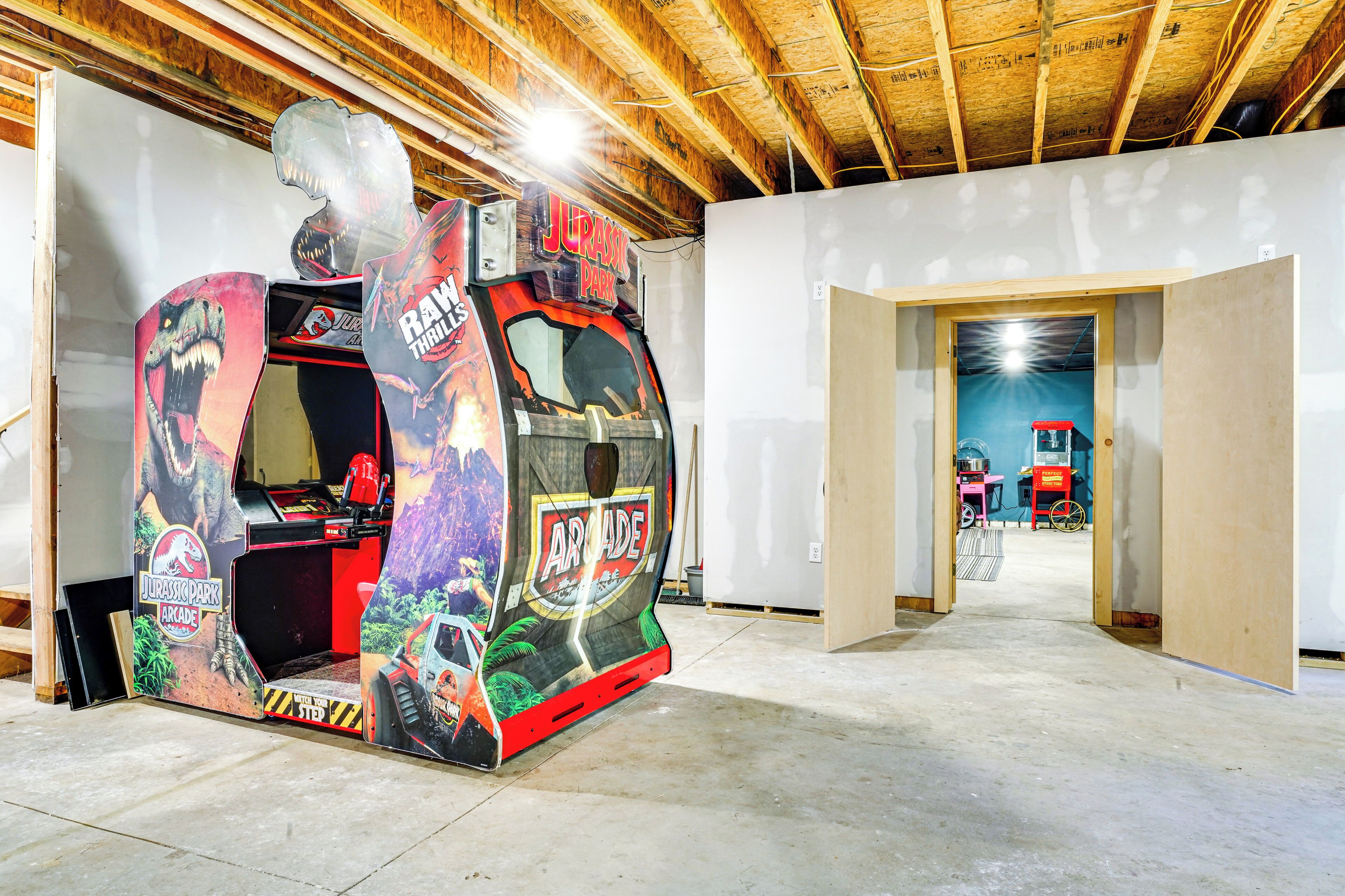 An arcade machine with a dinosaur theme in a room with exposed wooden beams and a concrete floor.