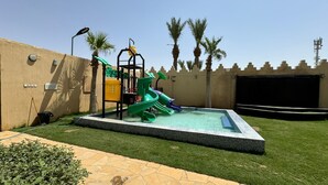 Un parque infantil con tobogán y estructuras de escalada, rodeado de una piscina y palmeras.