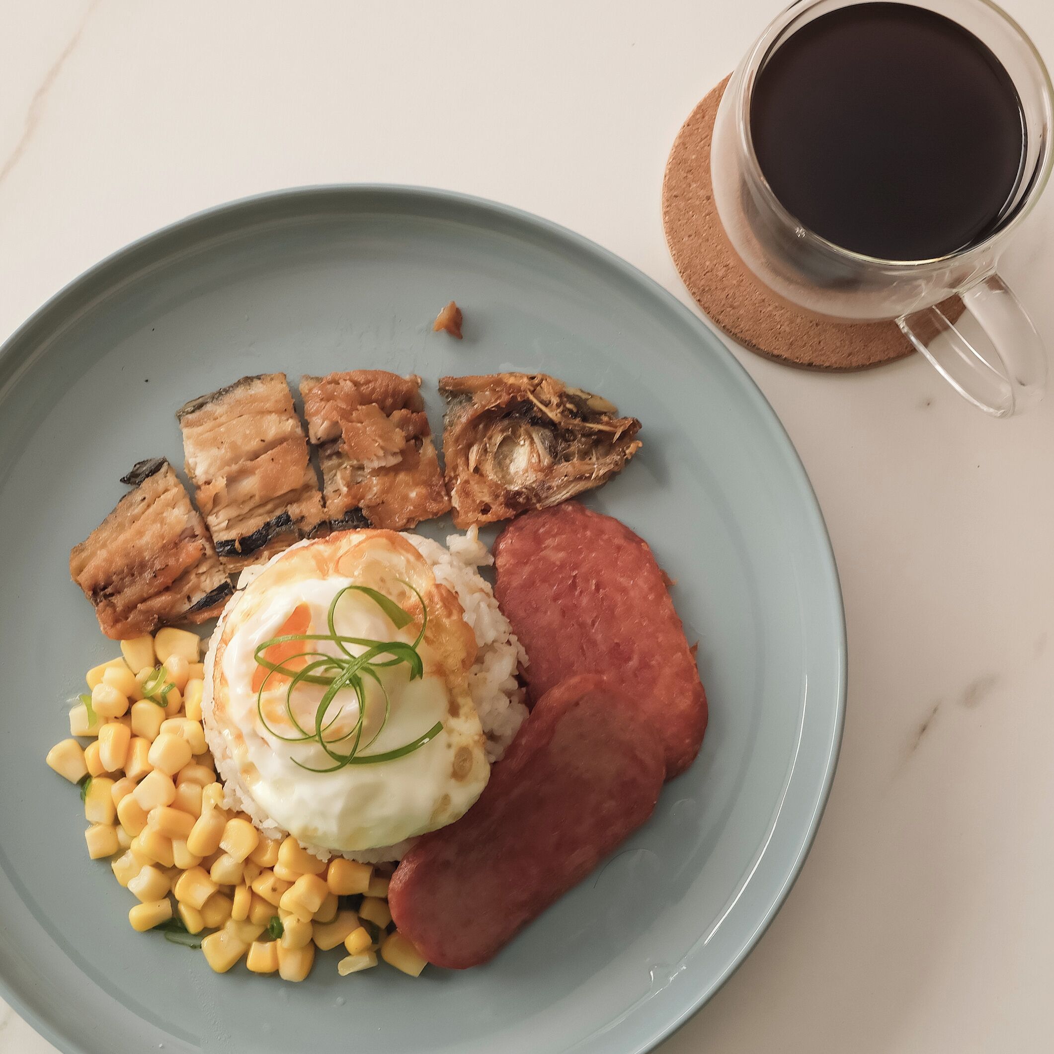 Un piatto con riso, mais, un uovo in camicia e fette di carne, accompagnato da una tazza di caffè nero su un sottobicchiere di sughero.