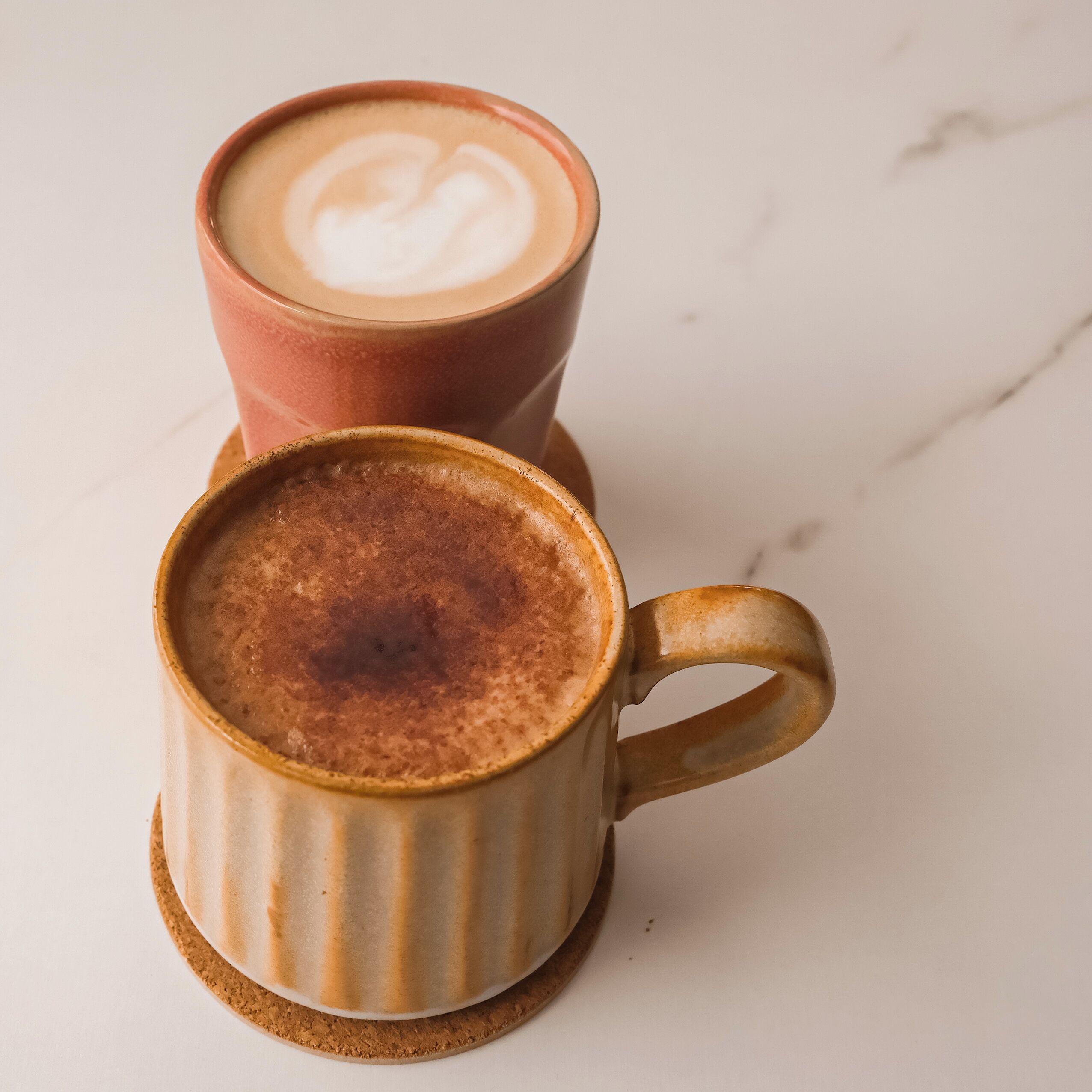 Deux tasses à café en céramique, dotées d’anses, dont l’une présente un motif de latte art, posées sur une surface blanche.