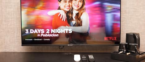Ein Flachbildfernseher zeigt '3 Days 2 Nights in Poblacion' auf Netflix, zusammen mit einer Kaffeemaschine und Fernbedienungen auf einem Holztisch.