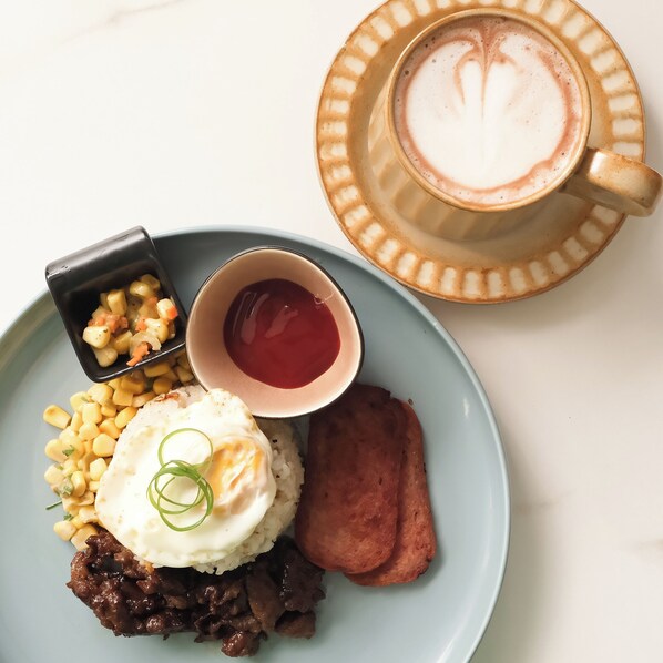 A plate with rice, a fried egg, corn, and a piece of meat, accompanied by a cup of coffee with latte art. - The Quarters by Rosey (Cabanatuan)