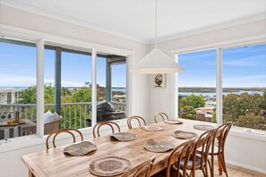 Un comedor con una mesa de madera preparada para seis personas, ventanas amplias que ofrecen vista al mar y follaje, y una lámpara colgante.