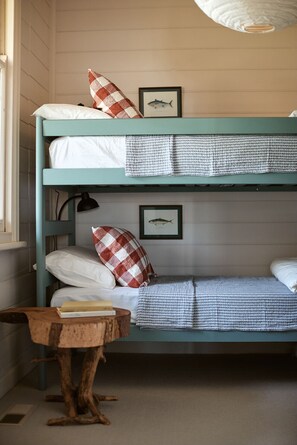 Uma cama beliche com dois níveis, cada um com um travesseiro e um quadro de um peixe. Há uma pequena mesa de madeira com um livro e um abajur no chão.