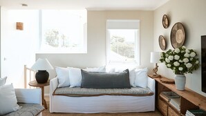 A cozy living room with a sofa, a side table with a lamp, a wooden bench, and a large window providing natural light. - Coastal Beach House | Panoramic Views and Stunning Sunsets (Ocean Grove)