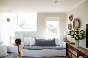A cozy living room with a sofa, a side table with a lamp, a wooden bench, and a large window providing natural light.