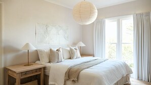 A bedroom with a large bed, two bedside lamps, a hanging light fixture, a window with curtains, and a wall art piece. - Coastal Beach House | Panoramic Views and Stunning Sunsets (Ocean Grove)
