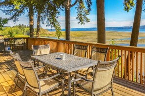 Eine Holzterrasse mit einem Metalltisch und -stühlen, mit Blick auf einen Strand und entfernte Berge.