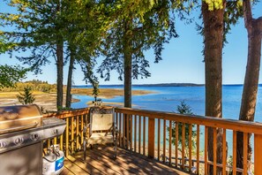 Eine Holzterrasse mit Grill und Stuhl, mit Blick auf ein Gewässer und Bäume.