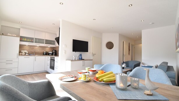 A modern kitchen with white cabinets, a flat-screen TV, and a dining area with a wooden table, chairs, and a fruit bowl. - Vacation apartment Molle (Norden)