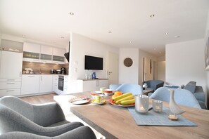 A modern kitchen with white cabinets, a flat-screen TV, and a dining area with a wooden table, chairs, and a fruit bowl. - Vacation apartment Molle (Norden)