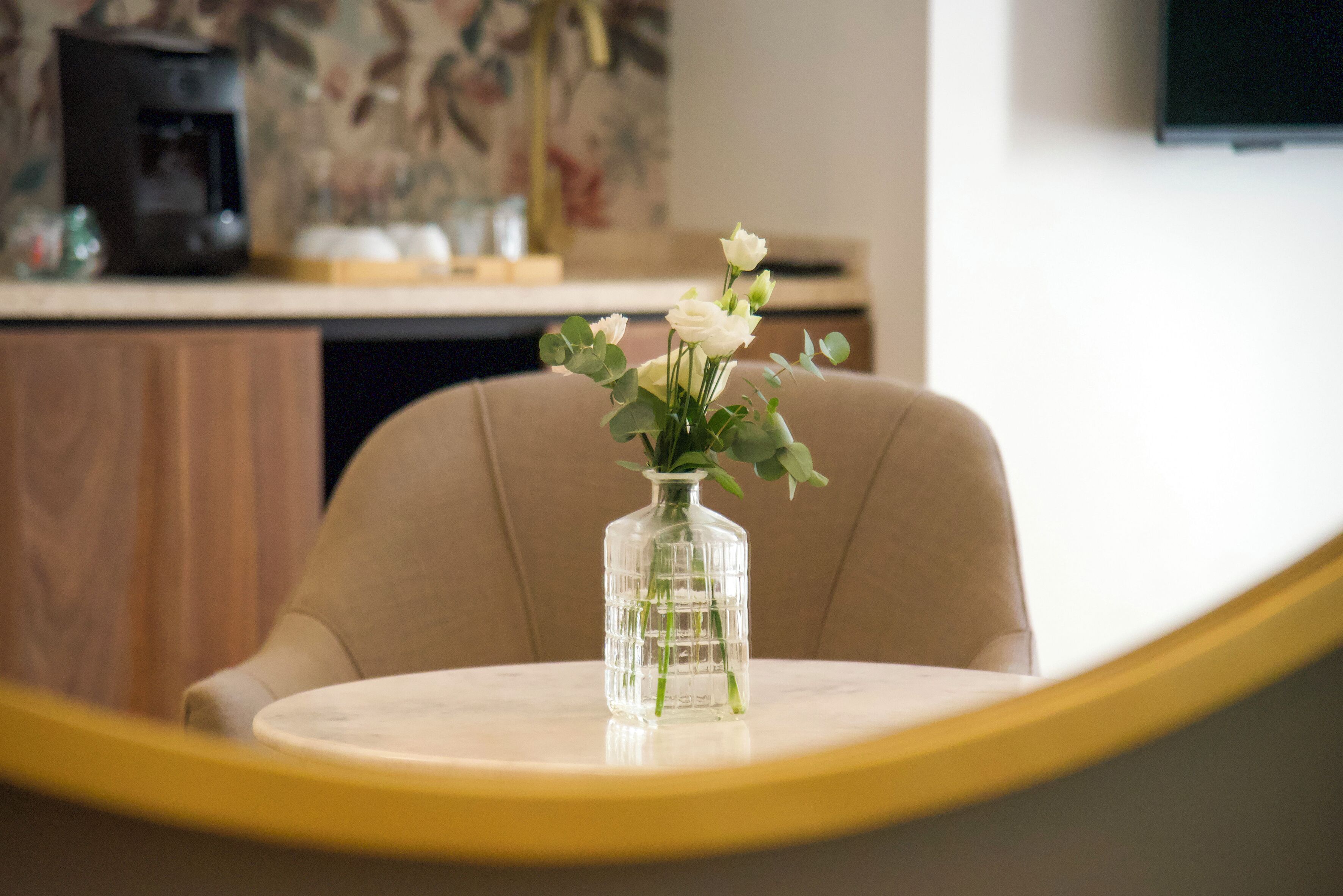 Un vaso di vetro trasparente con fiori bianchi su un tavolo rotondo in una stanza con un armadio in legno e un televisore appeso al muro.